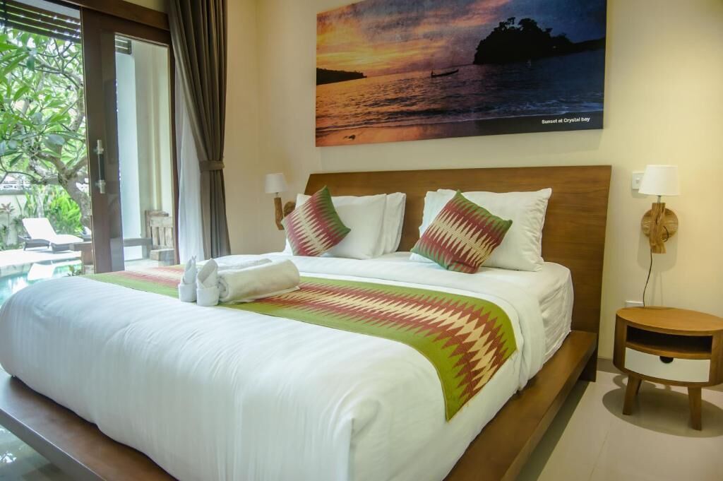 A hotel room with a large bed, a painting of a sunset, a view of the outdoors, and a small bedside table.