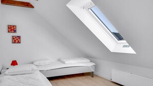 Une chambre avec un plafond en pente, un puits de lumière, un lit, une lampe rouge et deux tableaux encadrés.