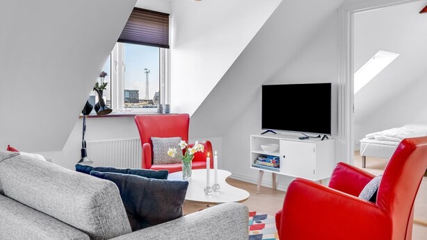 Un appartement moderne de style « loft » avec un plafond en pente, une grande fenêtre offrant une vue sur la ville, un téléviseur à écran plat et un coin salon confortable avec un fauteuil rouge et un canapé gris.