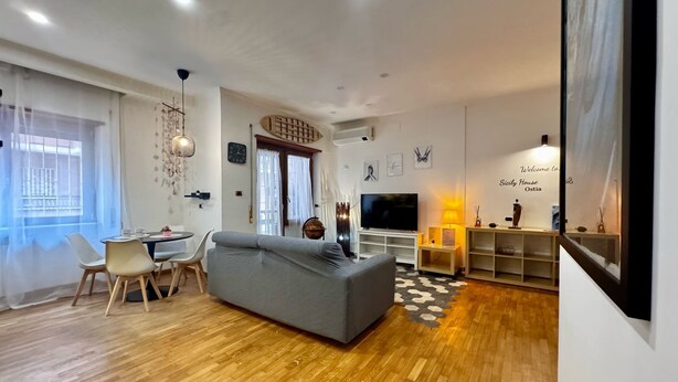 A modern living room with a grey sofa, wooden flooring, a dining area with chairs, a TV, and a bookshelf. - VACATION HOUSE IN OSTIA LEVANTE WITH 6 BEDS - ADJACENT LIDO NORD STATION (Lido di Ostia)
