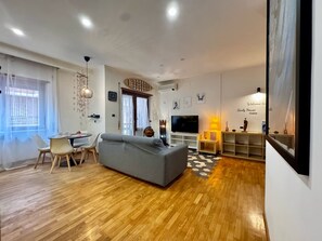 Un salon moderne avec un canapé gris, un plancher en bois, un coin repas avec des chaises, un téléviseur et une bibliothèque.