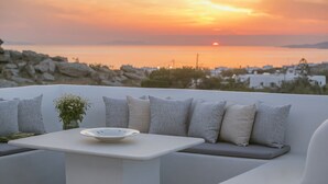 Een balkon met een witte tafel en zithoek, met uitzicht op een zonsondergang boven een kustlandschap.