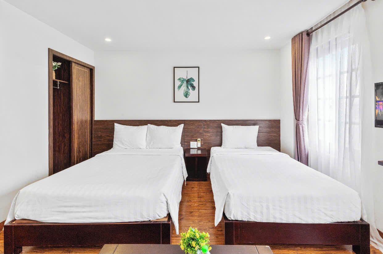 A hotel room with two single beds, a wooden headboard, a nightstand, a framed plant, and a window with curtains.