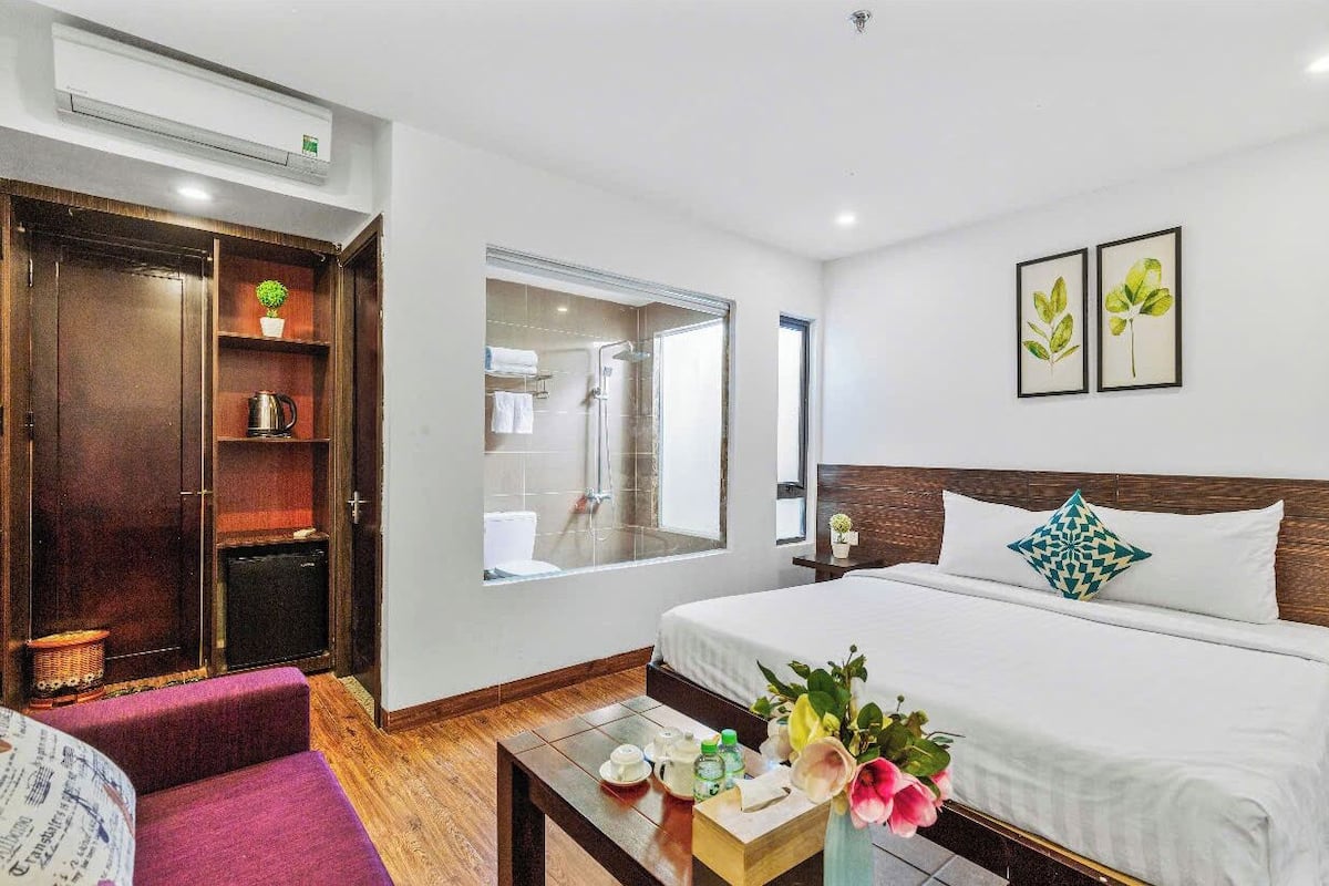A hotel room with a bed, a wooden wardrobe, a bathroom with a glass door, and a small table with a vase of flowers.