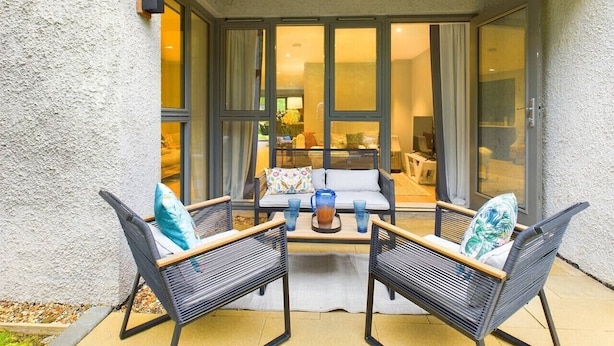 An outdoor seating area with two chairs and a table, a pergola overhead, and a view into the interior through glass doors. - Wild Roses | The Rowan Retreat · Lake District (Backbarrow)
