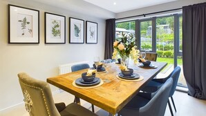 A dining area with a wooden table, chairs, framed botanical prints, and a view through large windows. - Wild Roses | The Rowan Retreat · Lake District (Backbarrow)