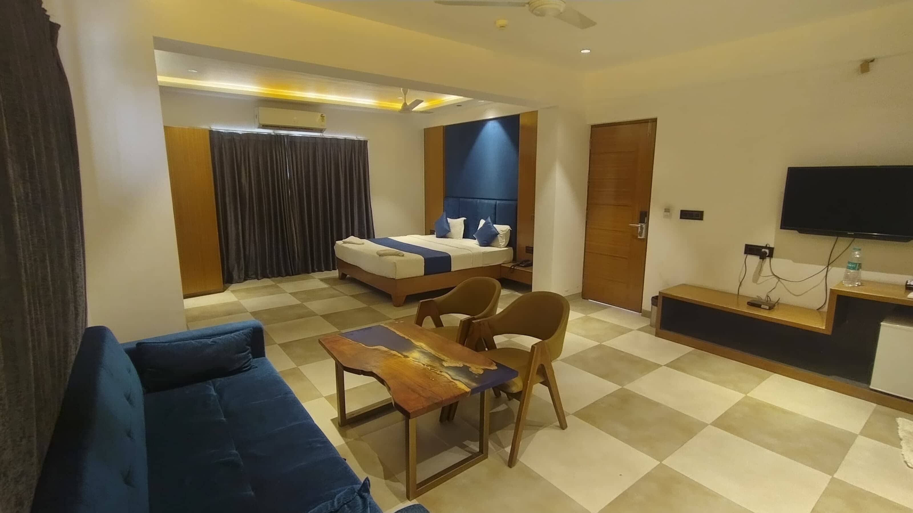 A modern hotel room with a checkered floor, a bed with blue and white bedding, a blue sofa, a wooden coffee table, and a TV on a wooden stand.