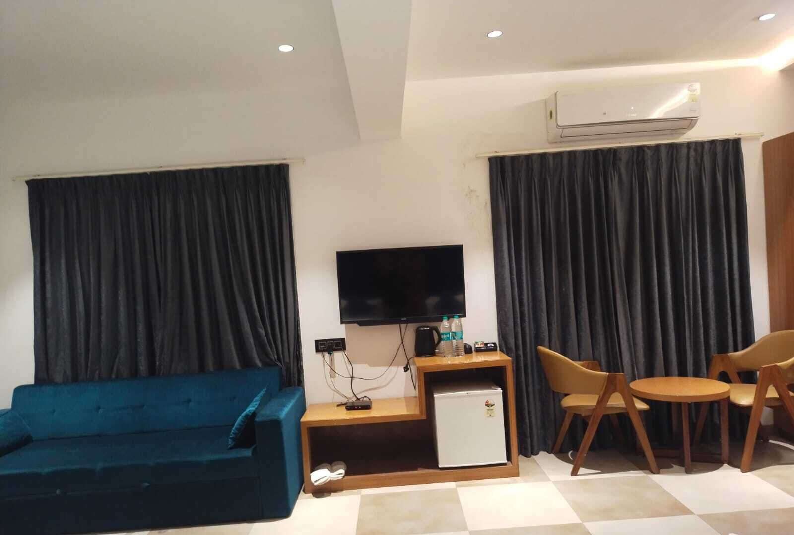 A modern hotel room with a blue sofa, a small wooden table with chairs, a flat-screen TV, a mini-fridge, and an air conditioning unit.