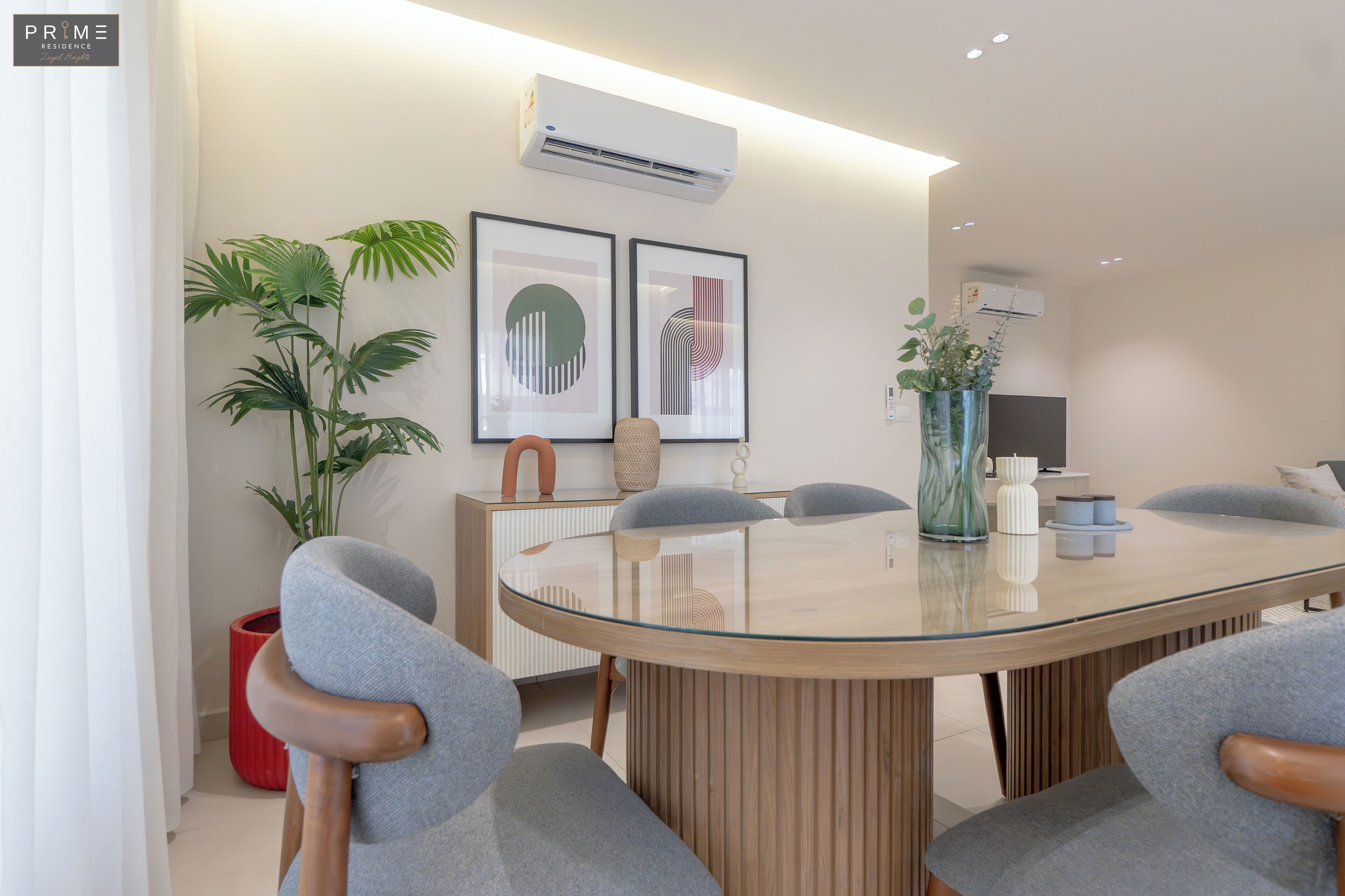 Un espace repas moderne avec une table à plateau en verre, des chaises à revêtement gris, une plante en pot et deux œuvres d’art encadrées accrochées au mur.