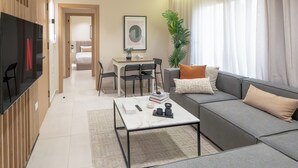 Ein modernes Wohnzimmer mit einem grauen Sofa, einem Couchtisch und einem Fernseher links. Im Hintergrund befindet sich ein Essbereich mit Tisch und Stühlen.