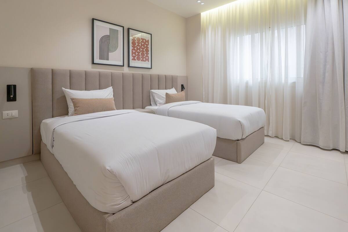 Ein Hotelzimmer mit zwei Betten, einem großen Fenster mit leichtem Vorhang und zwei gerahmten Kunstwerken an der Wand.