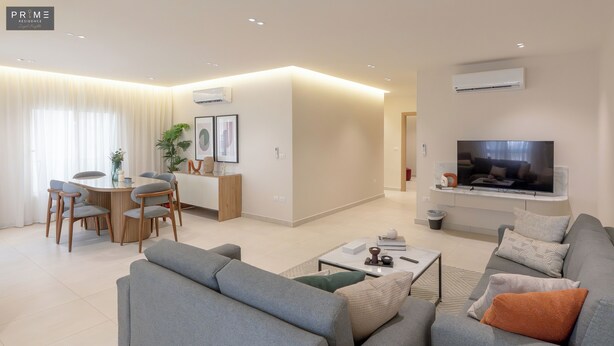 Ein modernes Wohnzimmer mit einem grauen Sofa, einem Couchtisch und einem an der Wand montierten Flachbildfernseher. Es gibt einen Essbereich mit Tisch und Stühlen sowie eine Küchenzeile mit Mikrowelle und Spüle.