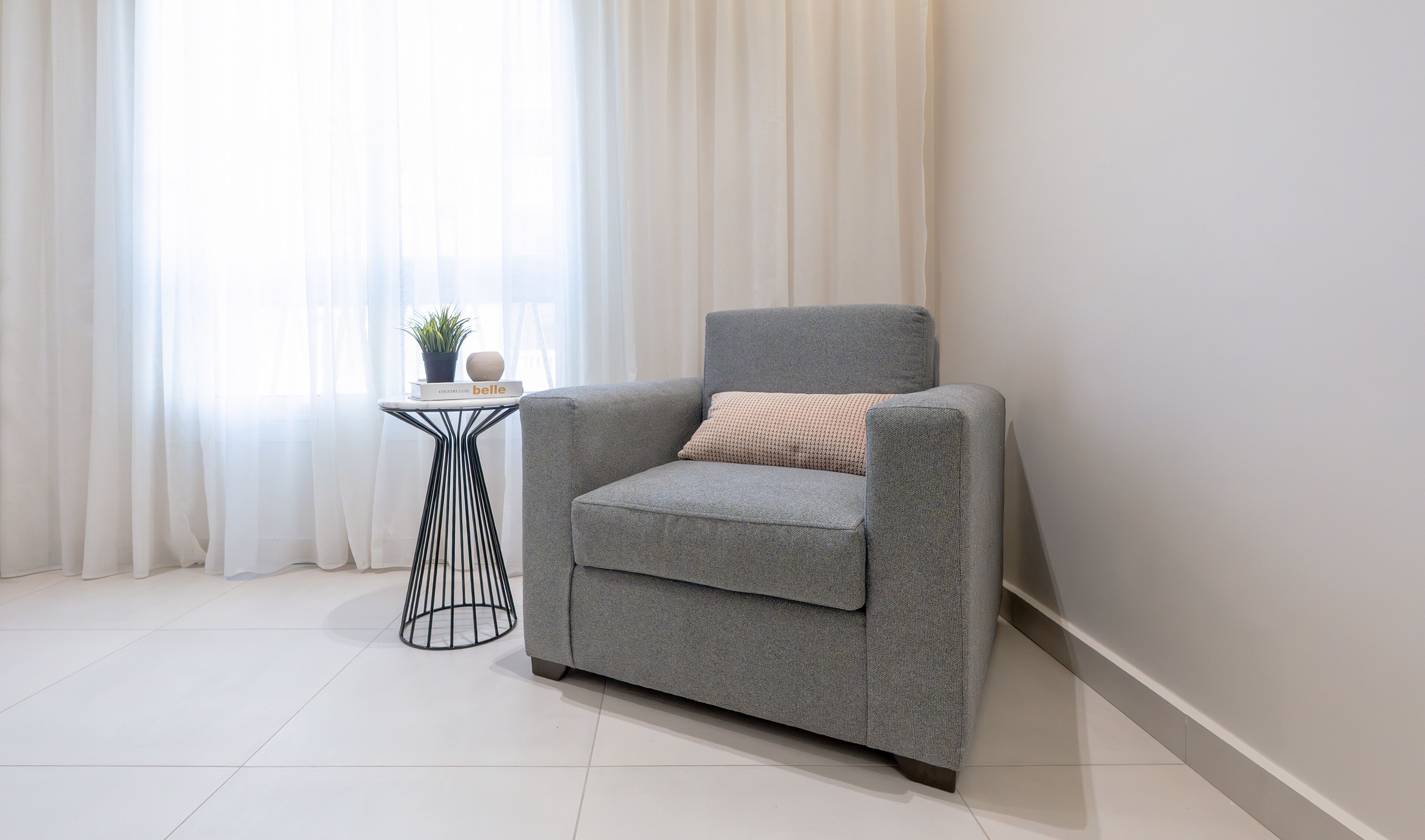 Un fauteuil gris avec un coussin, une petite table d’appoint avec une plante, et un livre intitulé « belle » dans une pièce avec des rideaux blancs et un sol carrelé.
