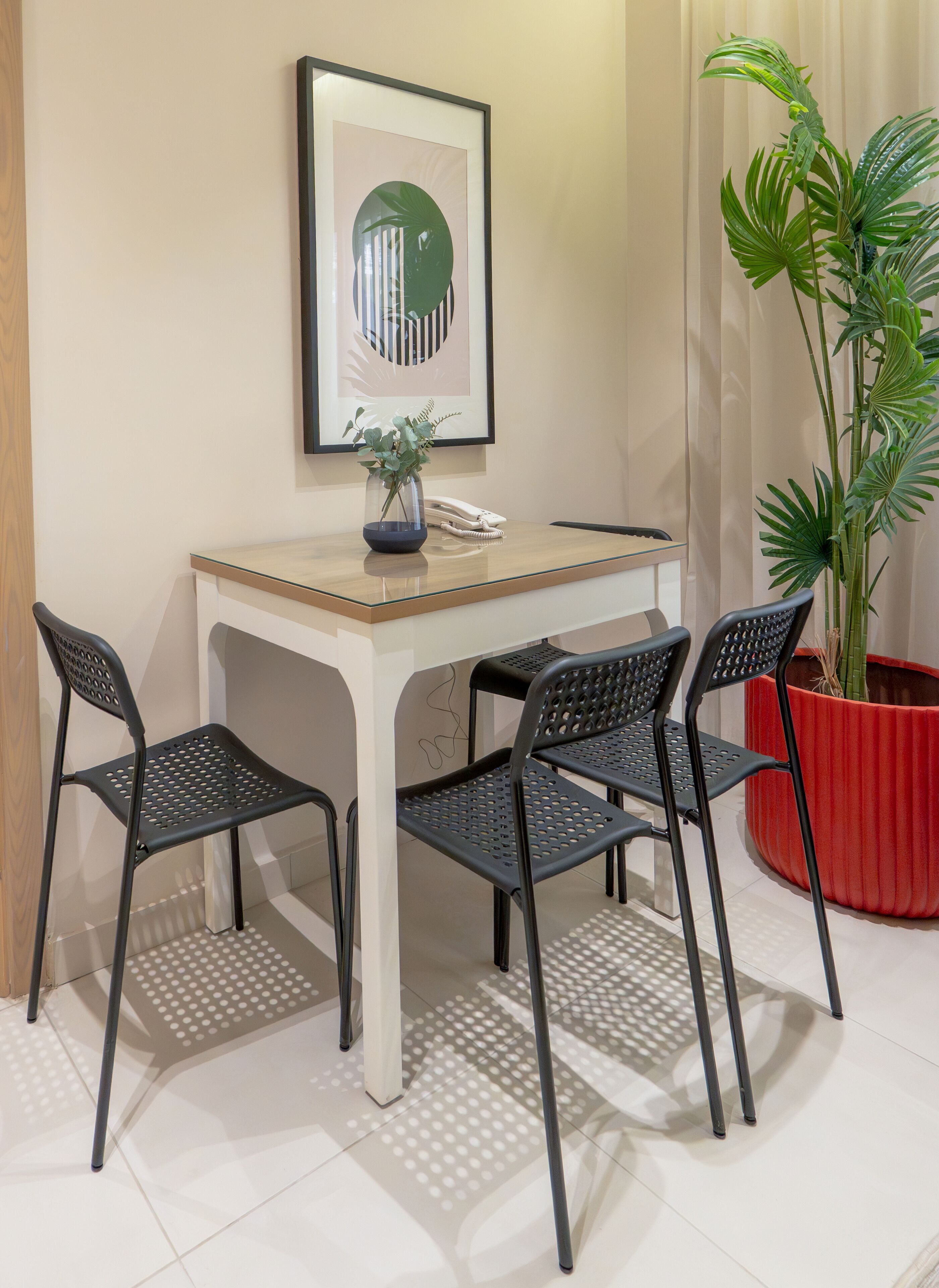 Habitación pequeña y moderna con una mesa de madera y cuatro sillas negras. Un cuadro enmarcado cuelga en la pared sobre la mesa, y hay una planta en maceta en la esquina.