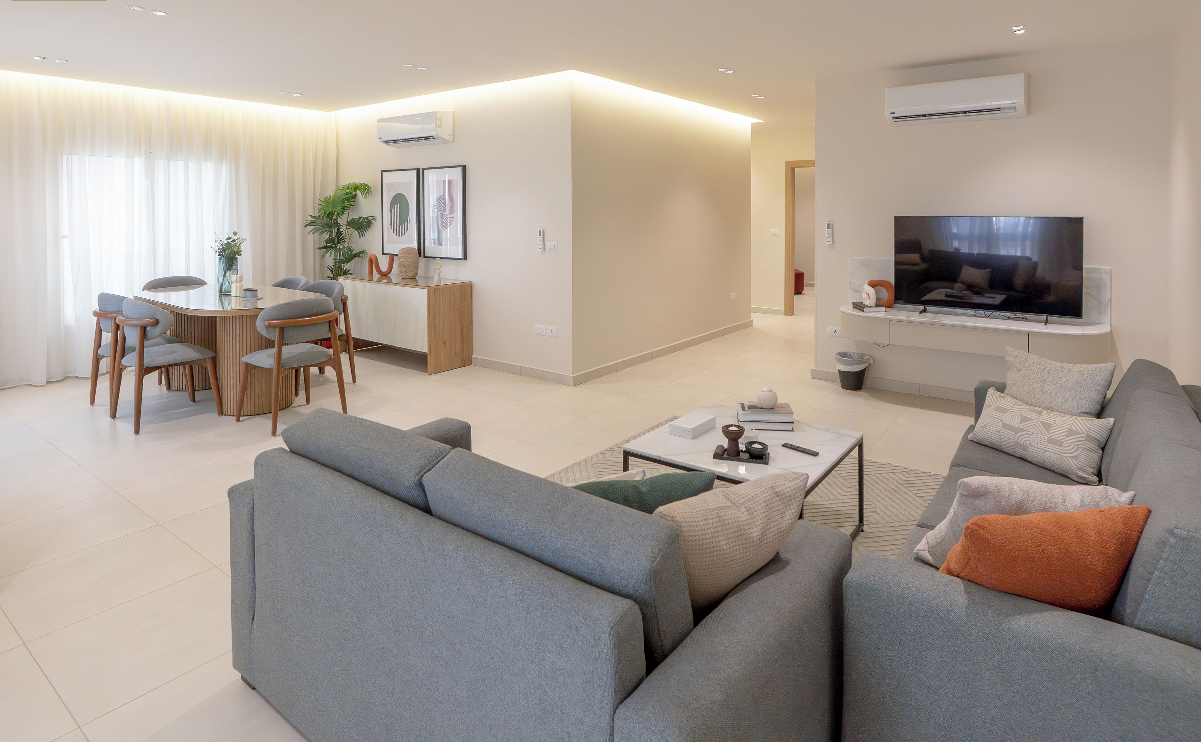 A modern living room with a grey sofa, a coffee table, and a flat-screen TV mounted on the wall. There is a dining area with a table and chairs, and a kitchen area with a microwave and a sink.