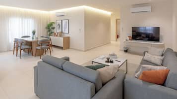 A modern living room with a grey sofa, a coffee table, and a flat-screen TV mounted on the wall. There is a dining area with a table and chairs, and a kitchen area with a microwave and a sink.
