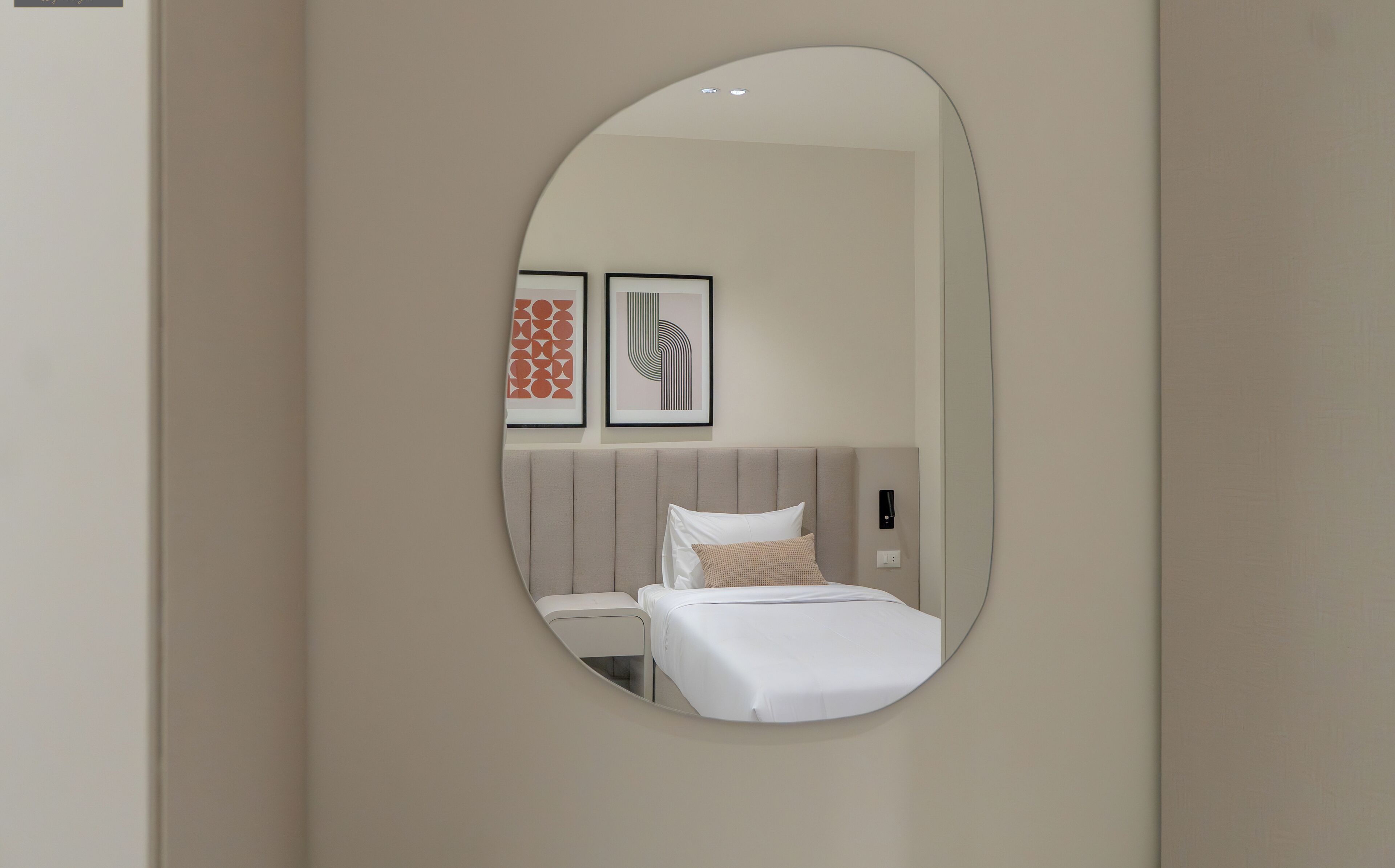 A modern hotel room with a round mirror, a bed with white linens, a headboard, and two framed artworks on the wall.