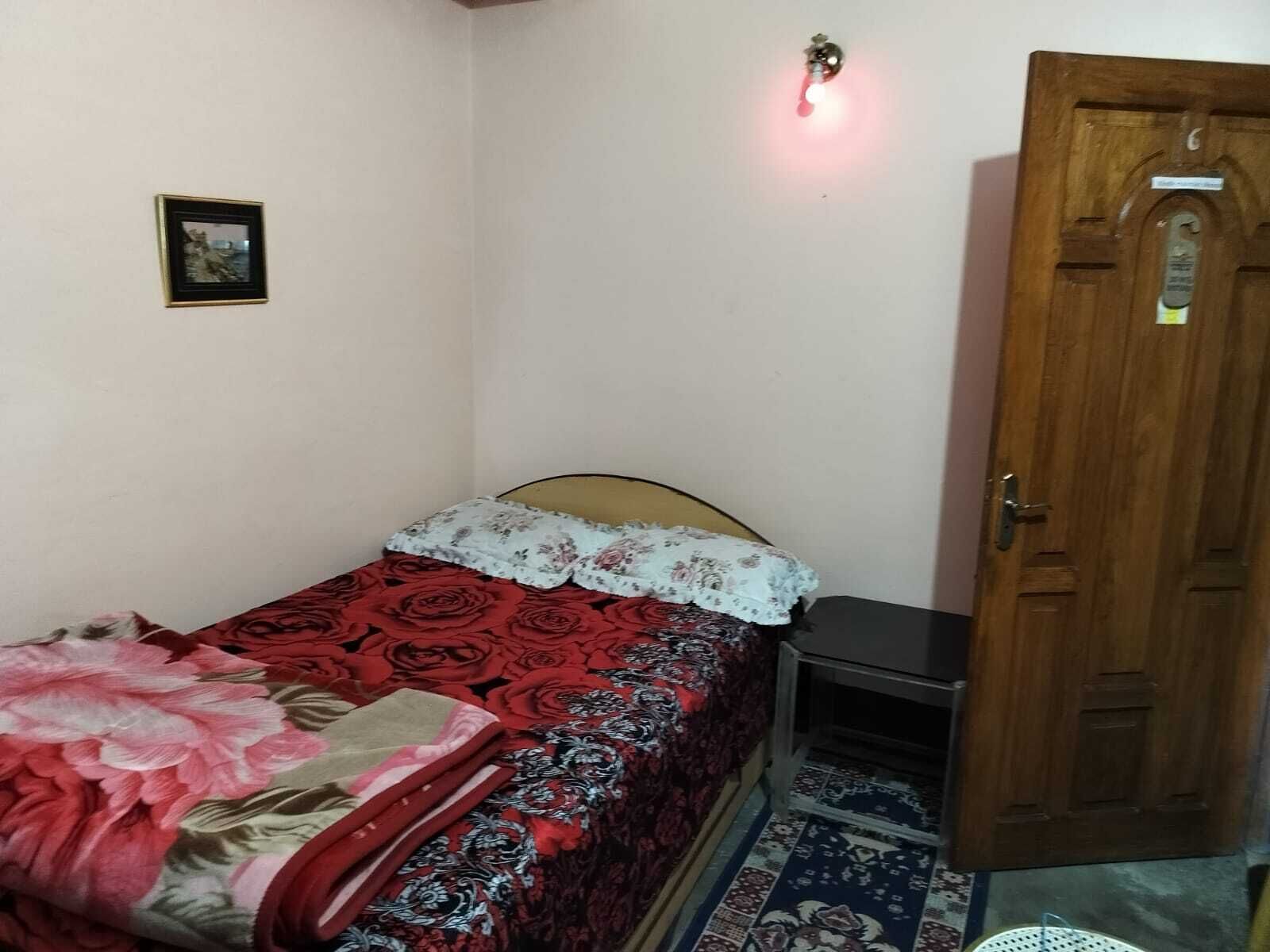 Habitació petita amb un llit individual, una tauleta de nit, una porta de fusta i una taula petita amb una làmpada.