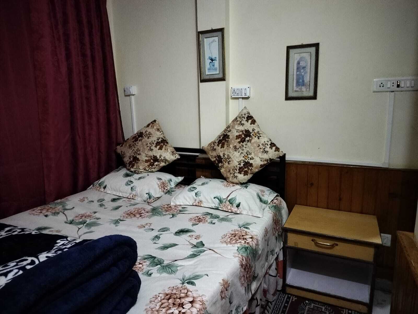 Een bed met bloemenbeddengoed, twee kussens, een houten nachtkastje met een lade en een rood gordijn.