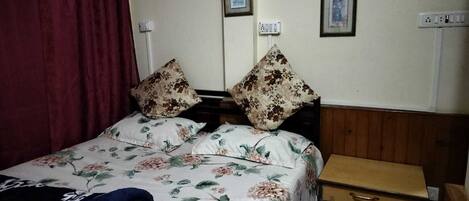 Een bed met bloemenbeddengoed, twee kussens, een houten nachtkastje met een lade en een rood gordijn.