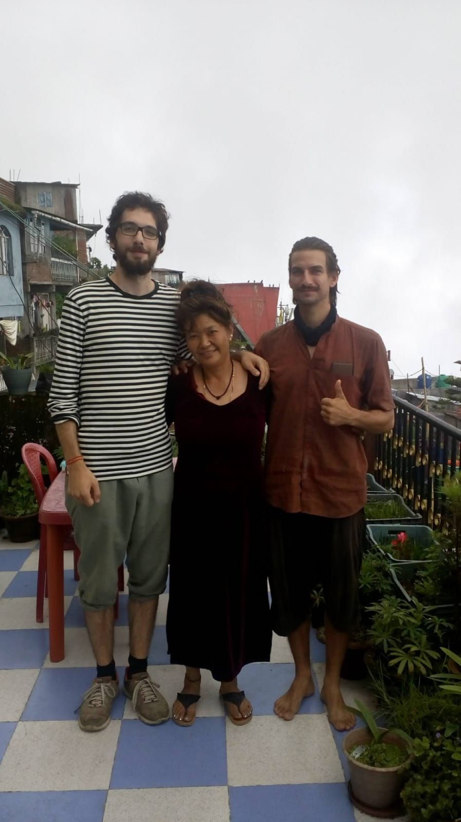 Three people standing on a checkered floor with potted plants and a red chair in the background.