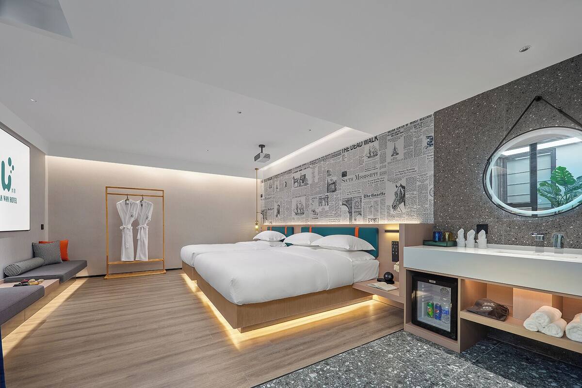 A modern hotel room with a large bed, a seating area, a vanity with a mirror, and a wall with newspaper clippings.