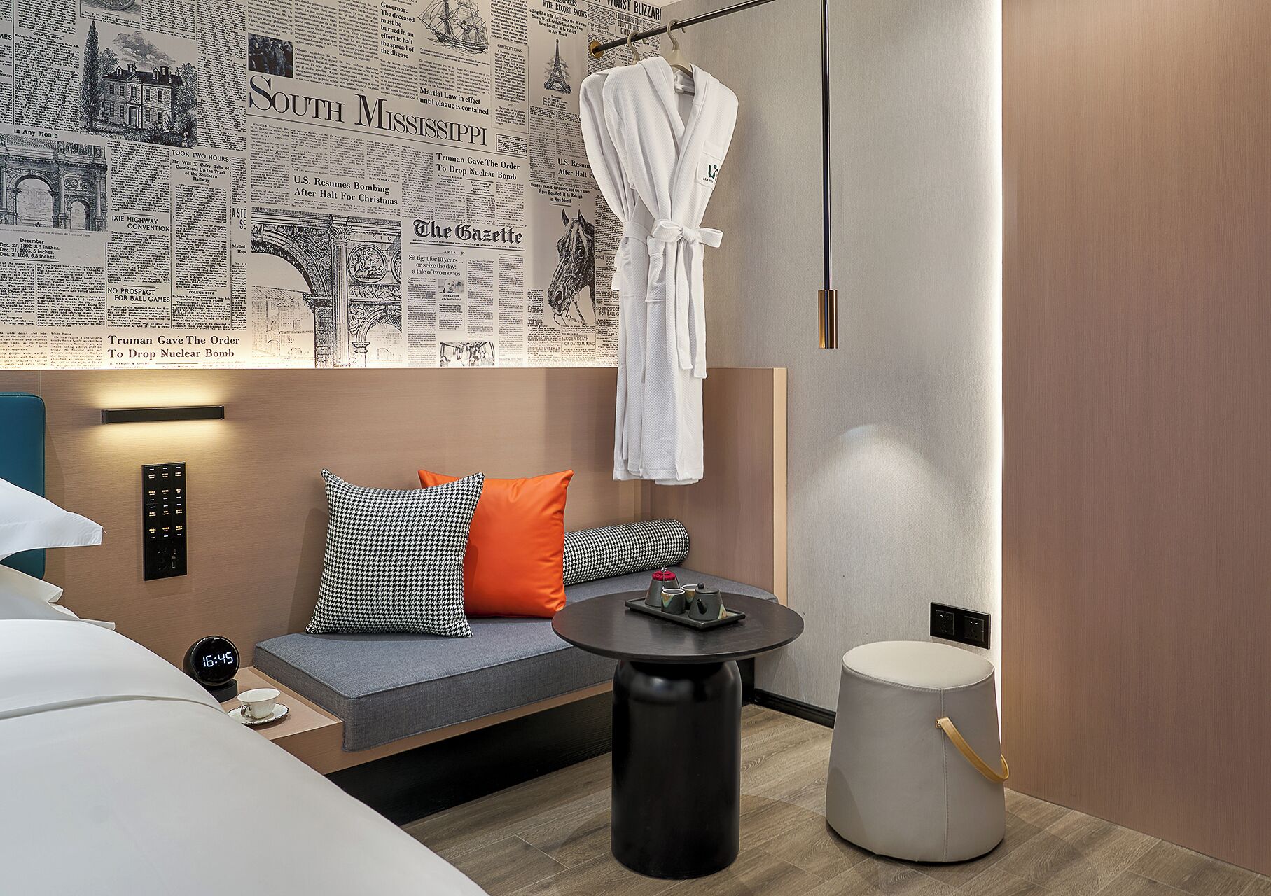 Una habitación de hotel moderna con un panel de pared cubierto de periódicos, un sofá con cojines estampados, una mesa redonda y una bata colgada en un gancho.
