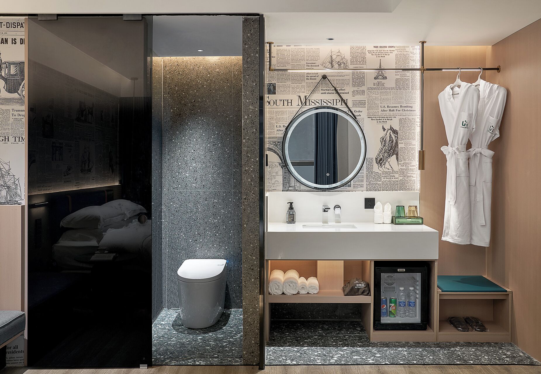 Un baño moderno con inodoro, lavamanos y espejo. Hay una bata colgada en un perchero, y un refrigerador pequeño con agua embotellada y artículos de aseo.