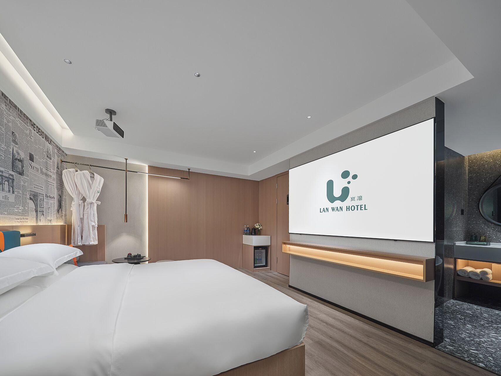 A hotel room with a bed, a TV screen displaying the hotel name, a robe hanging on a rack, and a small table with a lamp.