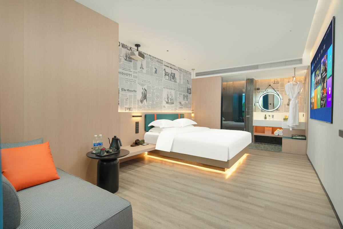 A modern hotel room with a large bed, a small desk, a chair, a sofa, a mirror, and a wall with a newspaper mural.