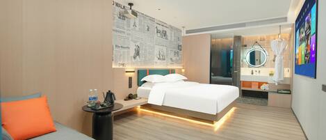 A modern hotel room with a large bed, a small desk, a chair, a sofa, a mirror, and a wall with a newspaper mural.