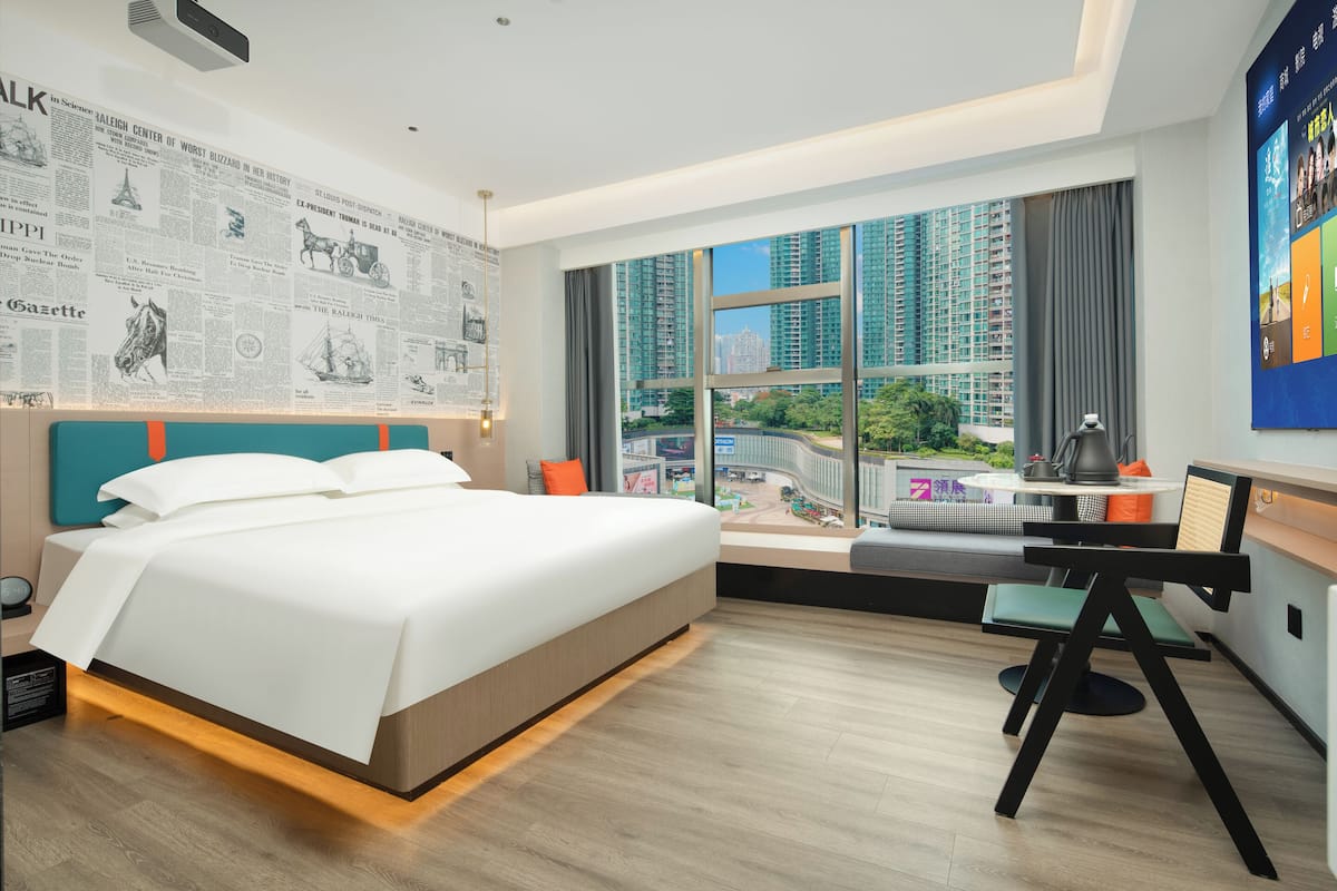A modern hotel room with a large bed, a desk, and a view of high-rise buildings.