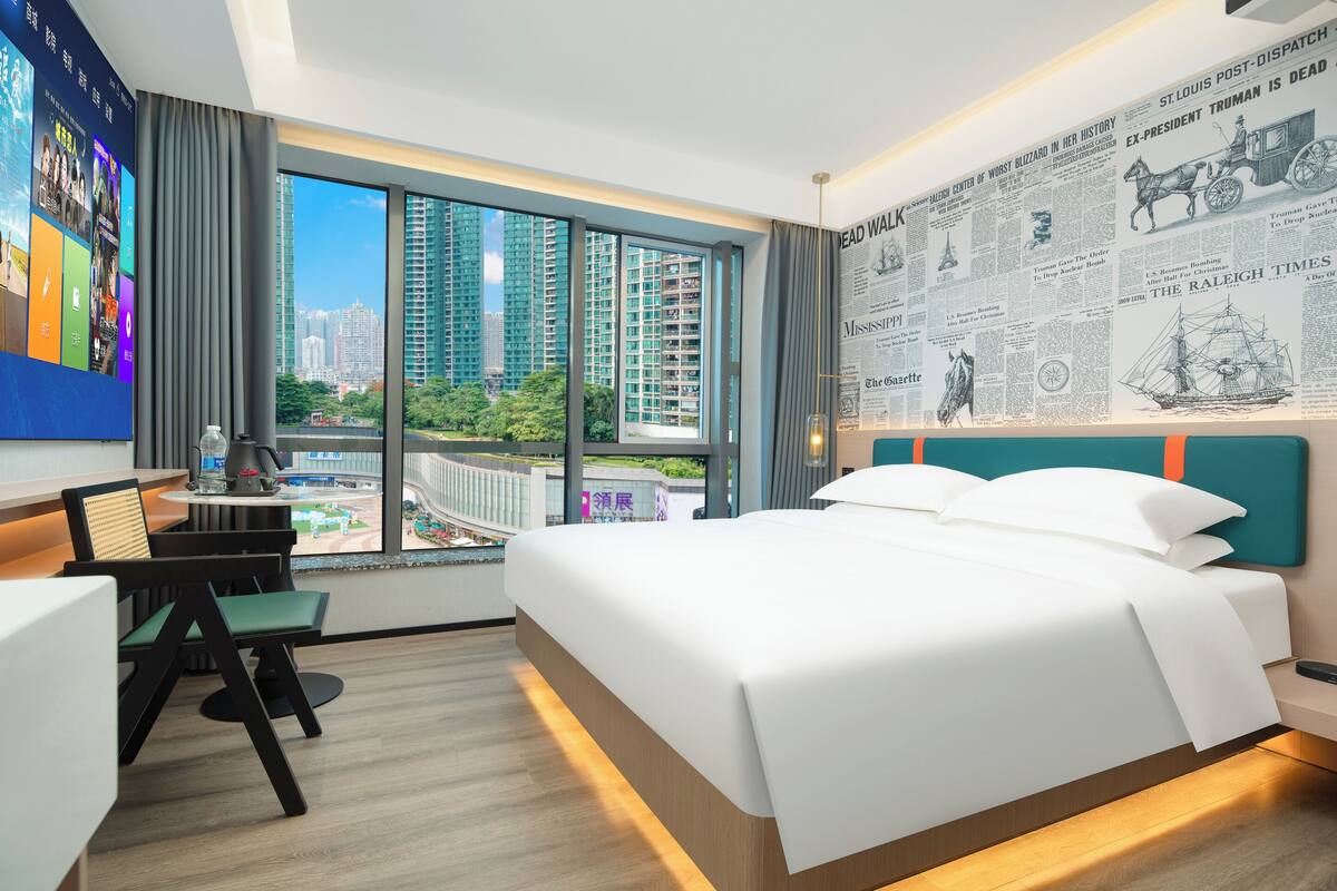 A hotel room with a large bed, a desk with a chair, a TV mounted on the wall, and a window with a city view.