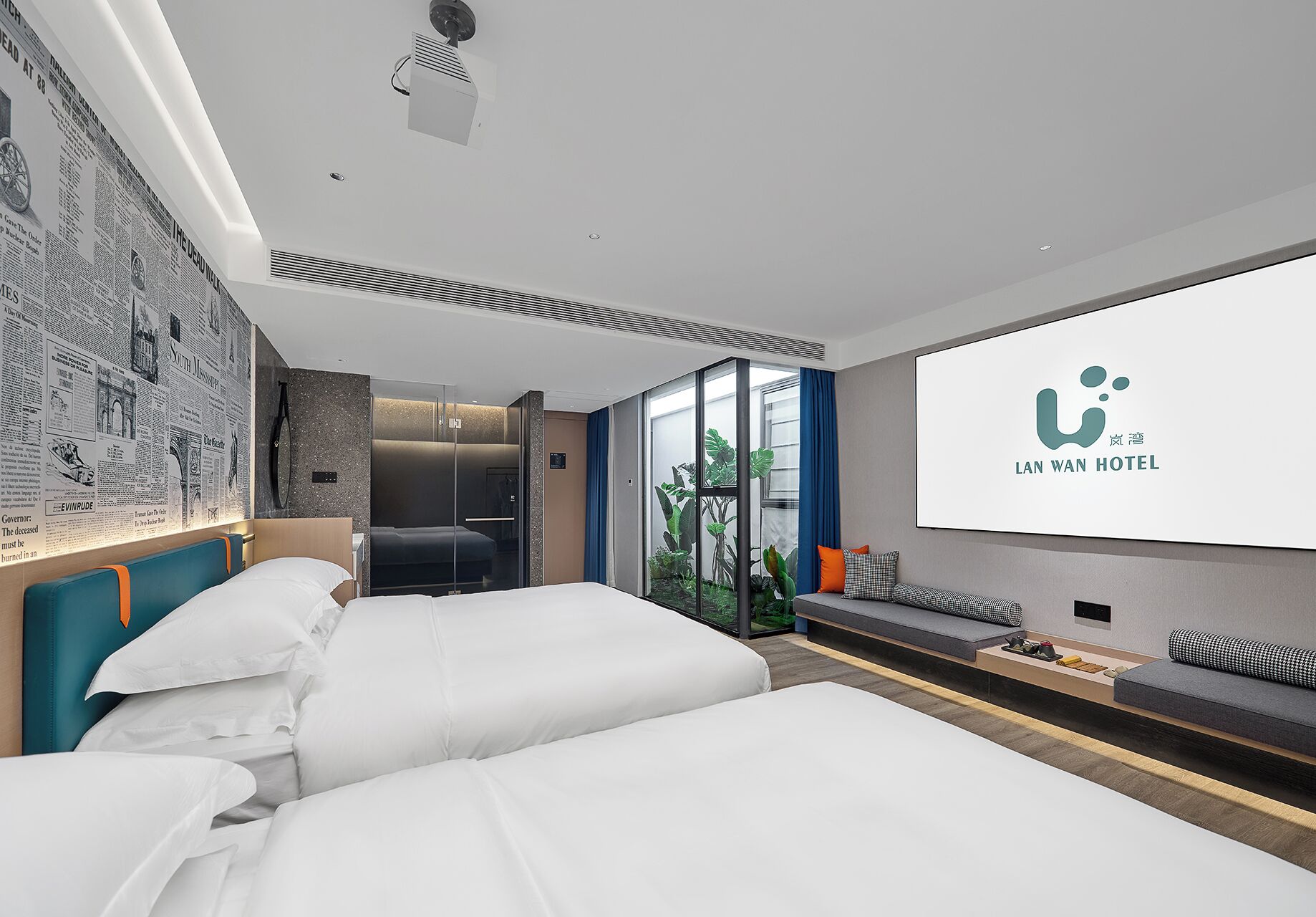 A modern hotel room with a large bed, a seating area with cushions, a glass partition, and a large screen displaying the hotel logo.
