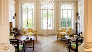 Une pièce spacieuse avec un plafond haut, des moulures décoratives et un lustre. On y trouve deux fauteuils et une table avec des chaises, une grande fenêtre donnant sur un paysage verdoyant, ainsi qu’une porte avec un panneau de verre.