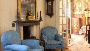 Une pièce avec deux fauteuils bleus, une cheminée ornée d’un miroir, une horloge et une vue sur un coin repas.