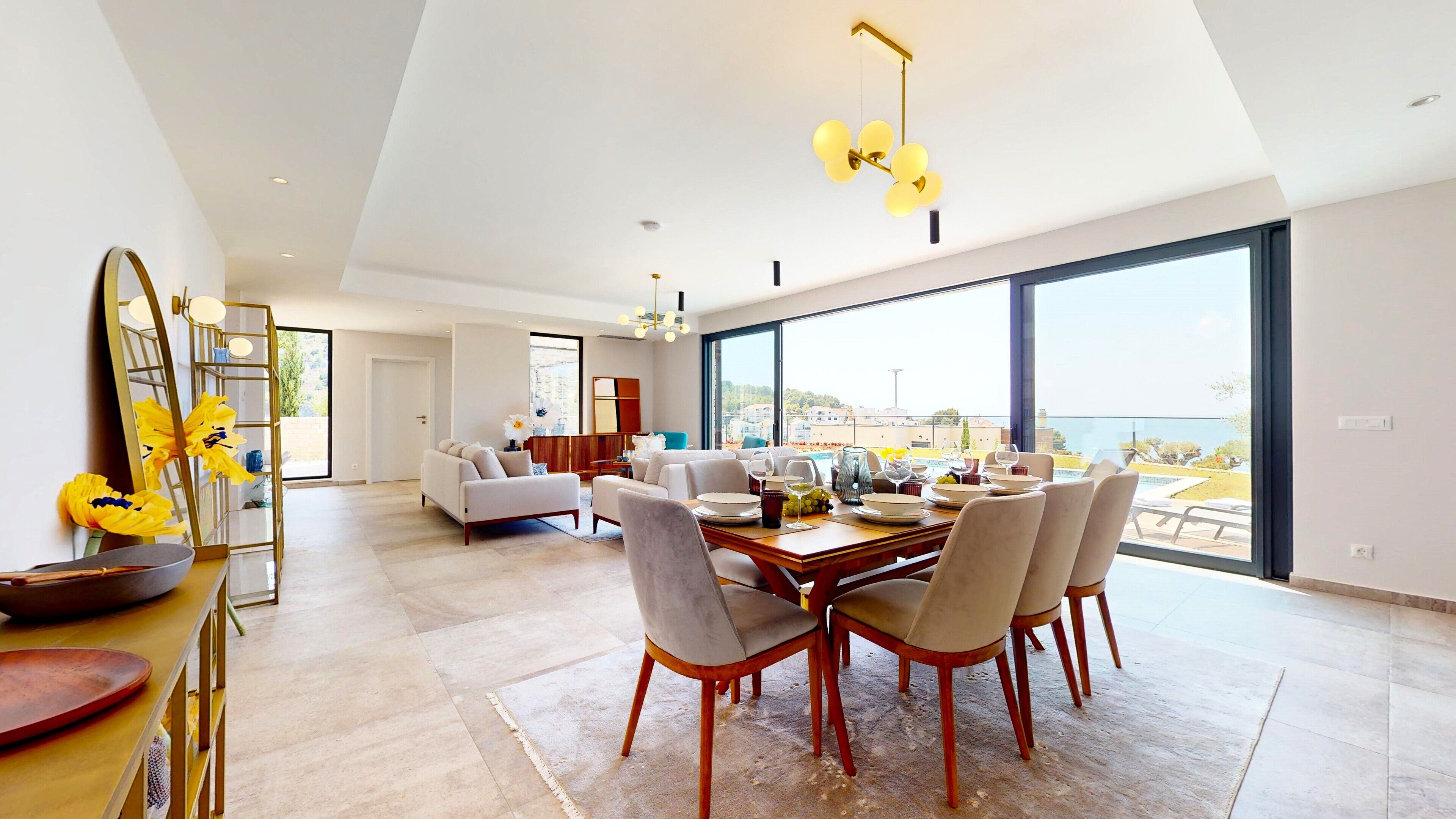 Une salle à manger spacieuse, avec une grande table dressée pour six personnes, un lustre moderne et une vue sur la mer grâce à de larges fenêtres.