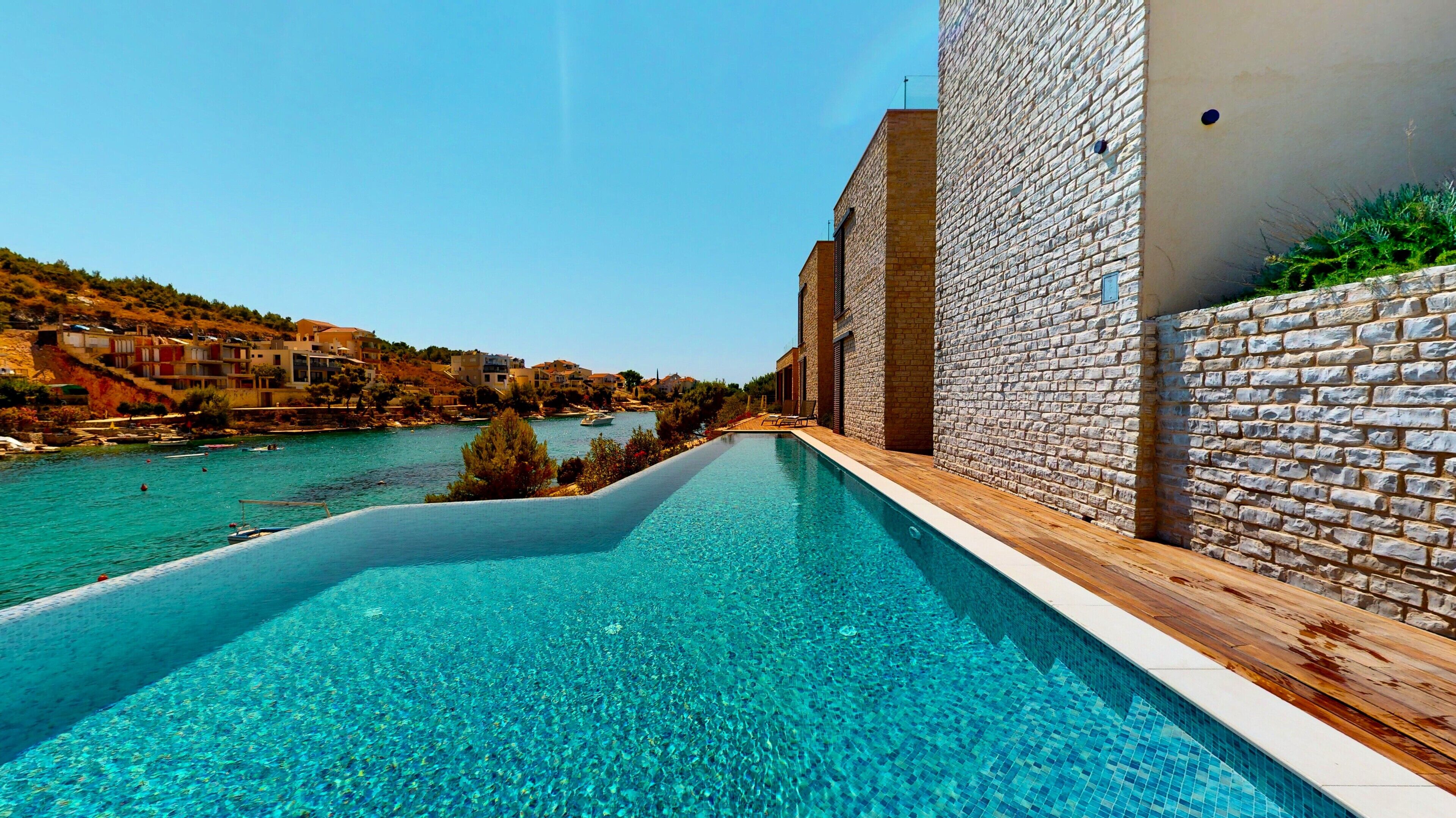 A swimming pool with a clear blue water, surrounded by a stone wall and wooden deck, overlooking a body of water and buildings in the background.