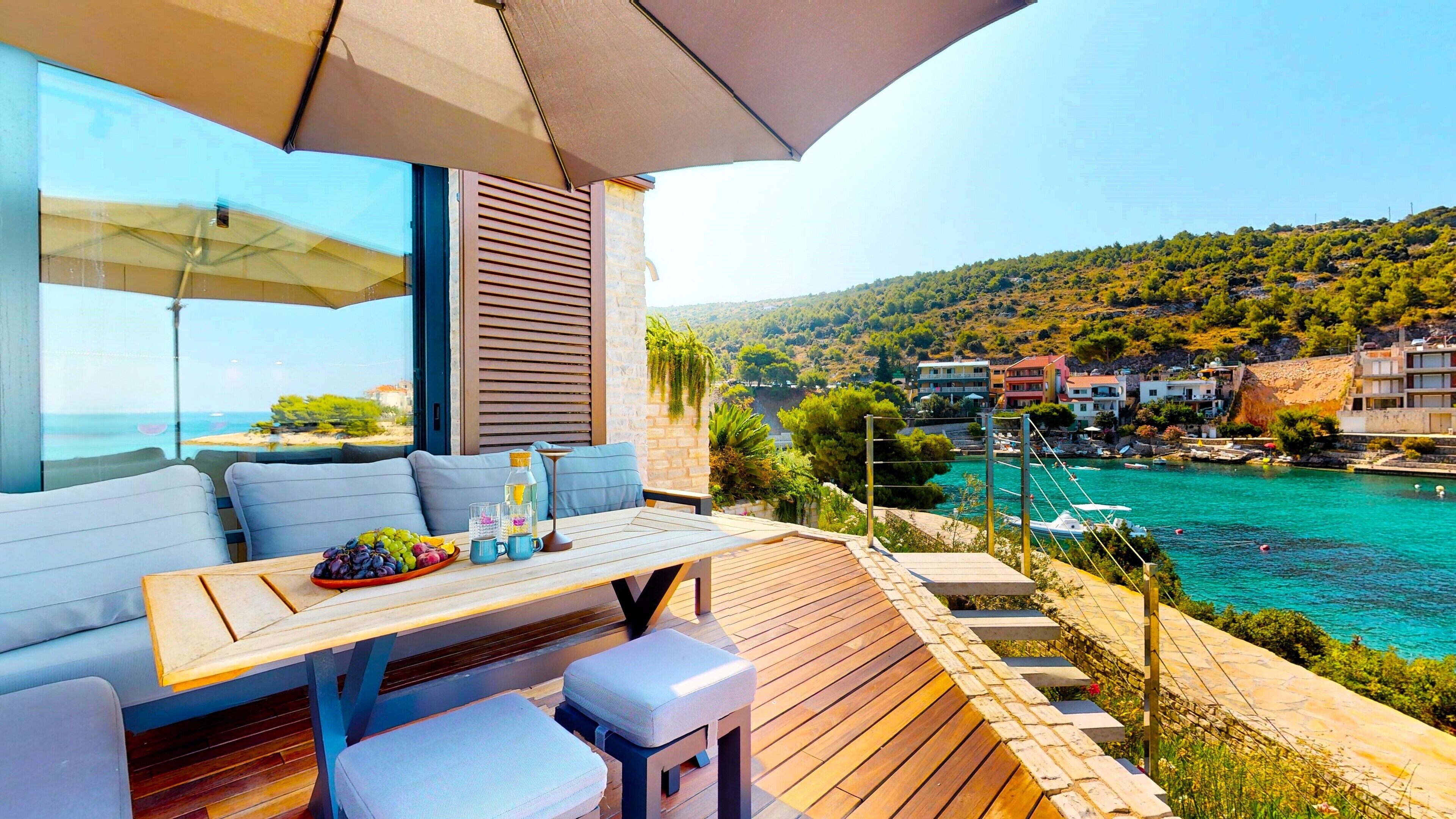 A balcony with a wooden table, seating, and a view of a coastal town and sea.