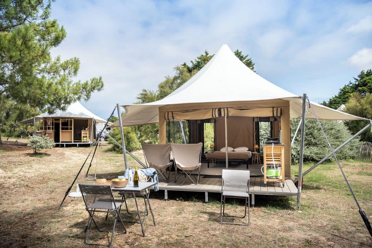 A tented accommodation with a dining area, outdoor seating, and a small table with a bottle and glasses.