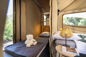 A cozy interior with a bed, a small table with a lamp, a teddy bear, and a window with a view of greenery. - Huttopia Ars en Ré  (Ars-en-Ré)