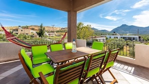 A balcony with a wooden table and green chairs, a hammock, and a view of a mountainous landscape.