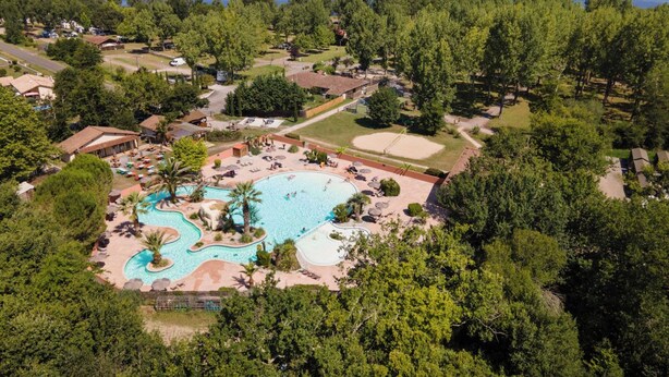 An aerial view of a resort with a large swimming pool, surrounded by lush greenery and a lake in the background.