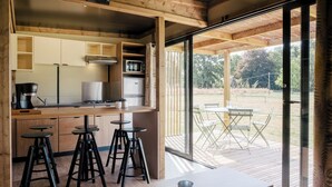 A modern kitchen with wooden cabinets, a built-in microwave, and a coffee maker. There are three stools and a table with a metal cup. The kitchen opens to a deck with a table and chairs.