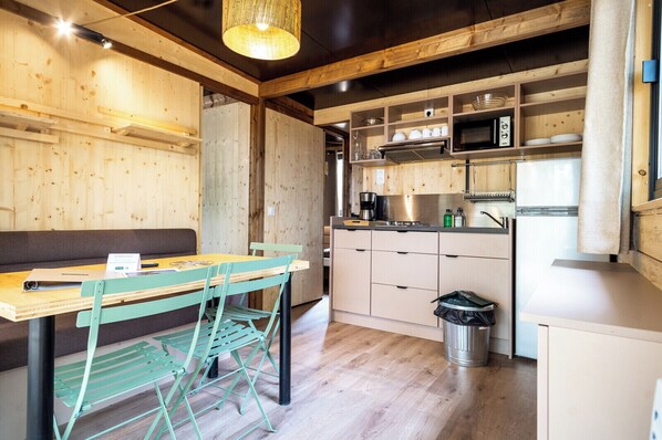 A compact kitchen with wooden cabinetry, a sink, and a microwave. There is a dining area with a table and chairs. - Huttopia Lac d'Aureilhan - Mimizan (Aureilhan)
