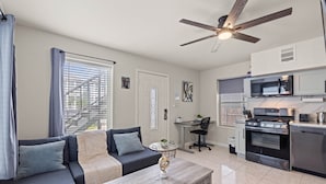 A modern living room with a sofa, coffee table, and a ceiling fan. Adjacent is a kitchen with a stove, microwave, and cabinets.