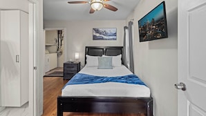 A bedroom with a bed, a ceiling fan, a painting, a TV, and a bathroom in the background.