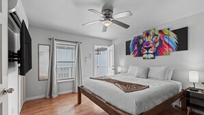 Een slaapkamer met een bed, een plafondventilator, een raam met gordijnen en een kunstwerk aan de muur.