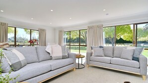 A spacious living room with two grey sofas, a small round table, and a large window offering a view of a garden. - Relaxing Rural Getaway – Near Kaiapoi Lakes, Room to Roam (Kaiapoi)
