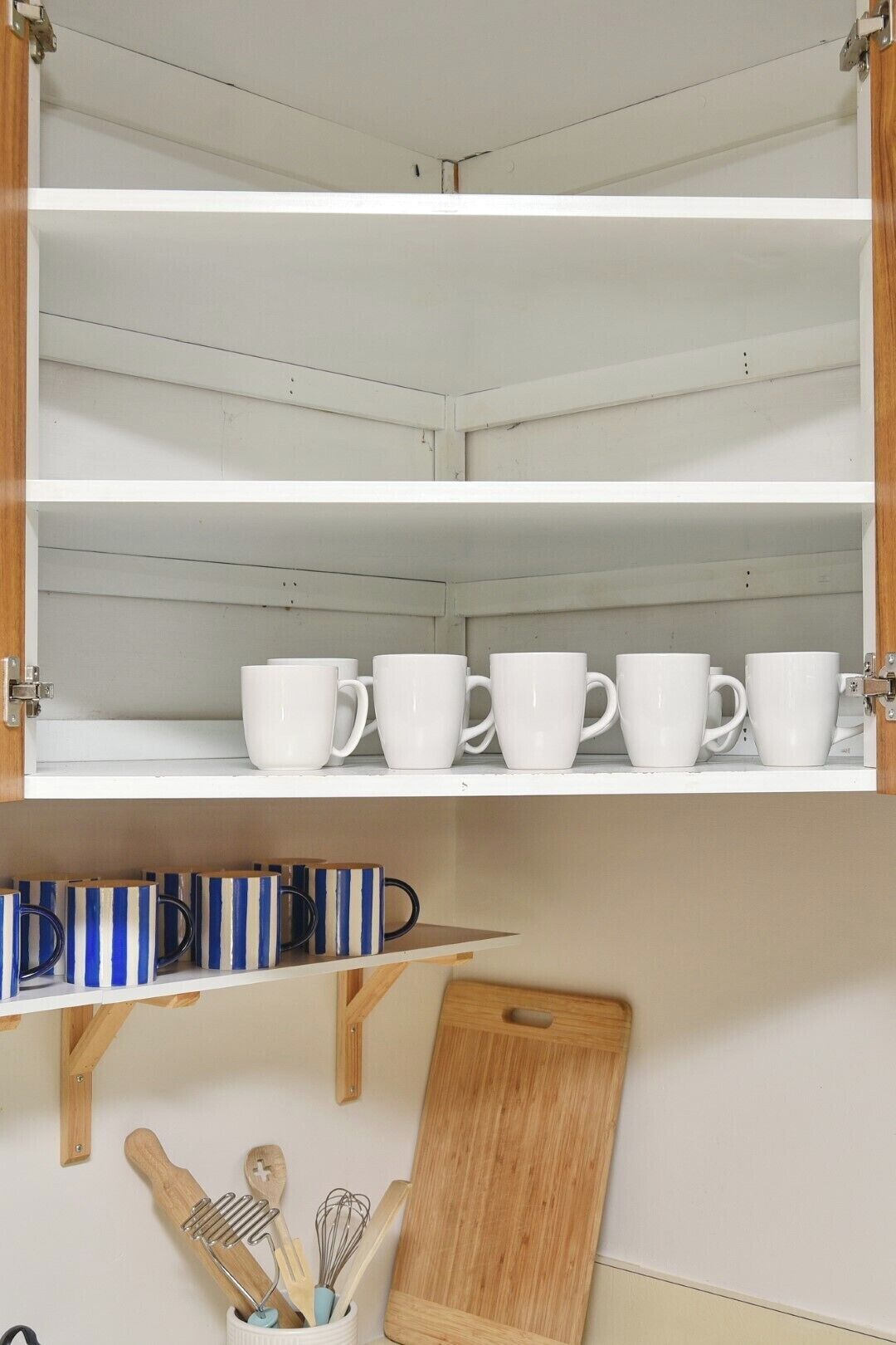 A kitchen with white cabinets, a wooden cutting board, and a shelf with striped mugs.